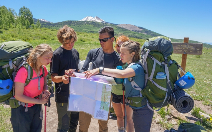 A group of people wearing backpacks examine a map. In the distance, there is a snow-capped mountain. 